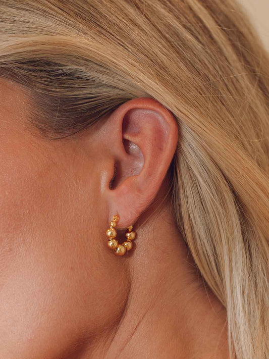 A close-up of a model's ear wearing a small gold hoop earring adorned with clustered round gold beads.
