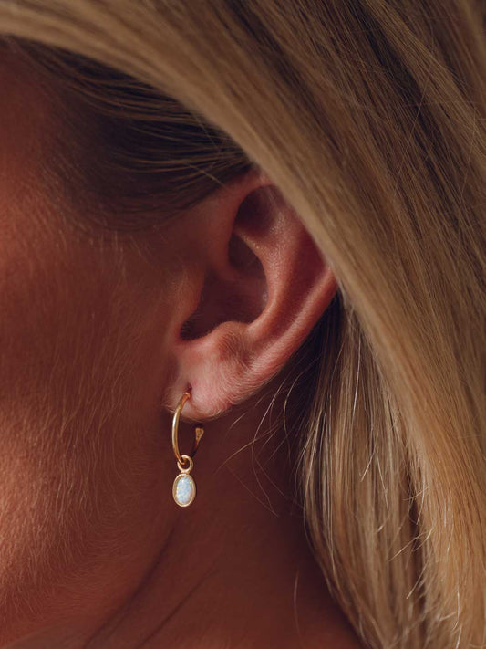 A close-up side profile of a woman's ear wearing a delicate gold hoop earring with a small opal teardrop stone.