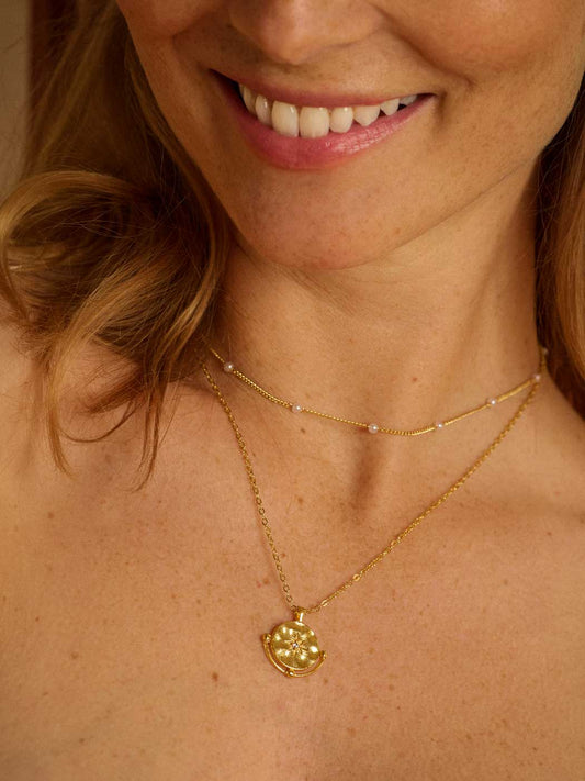 A close-up of a woman wearing a delicate gold necklace with a small gold pendant, layered over a shorter pearl choker.