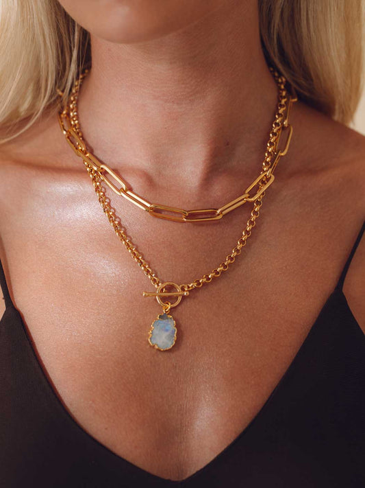A close-up of a model wearing a layered gold necklace set. The set includes a chunky paperclip chain and a delicate chain with a toggle clasp and a teardrop-shaped light blue stone pendant.