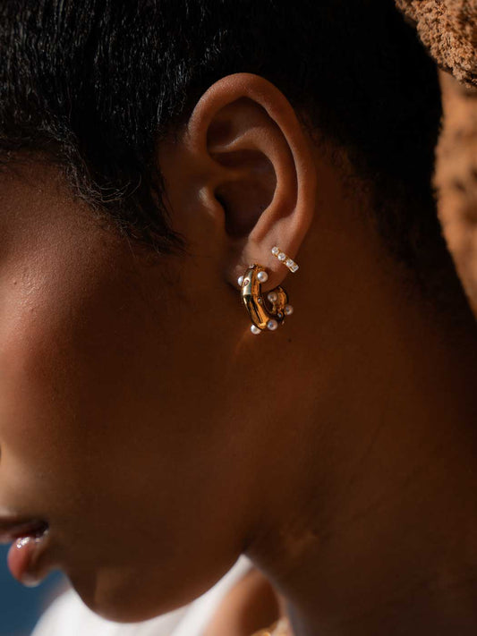 Close-up of a person's ear wearing a pair of gold huggie hoop earrings adorned with small white pearls, set against dark skin and short black hair.