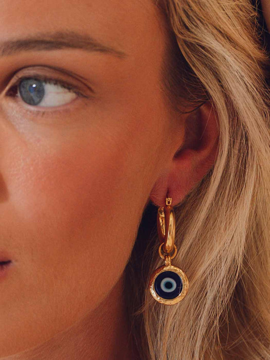 Close-up of a person wearing gold hoop earrings with blue evil eye charms.