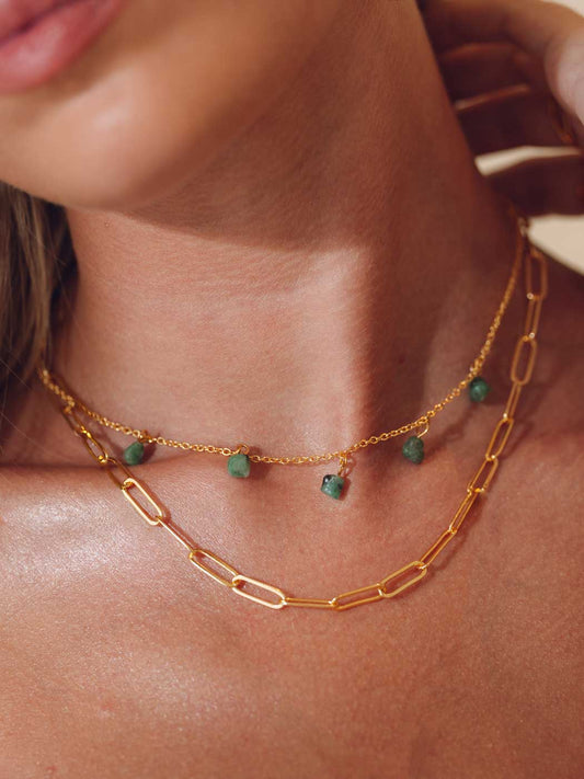 A close-up of a model's neck and upper chest wearing two layered gold necklaces: a paperclip chain and a delicate chain with small green bead accents.