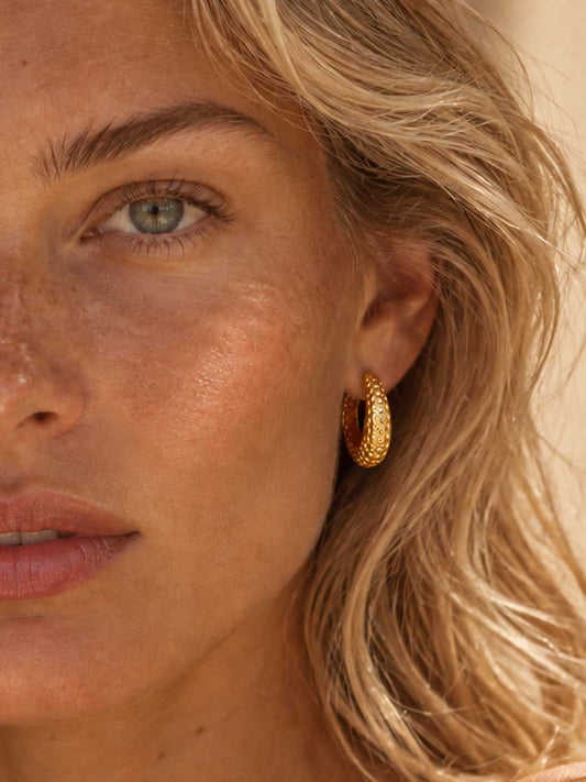Close-up of a woman wearing a textured gold hoop earring, showing the earring on her ear against her blonde hair and warm skin tones.