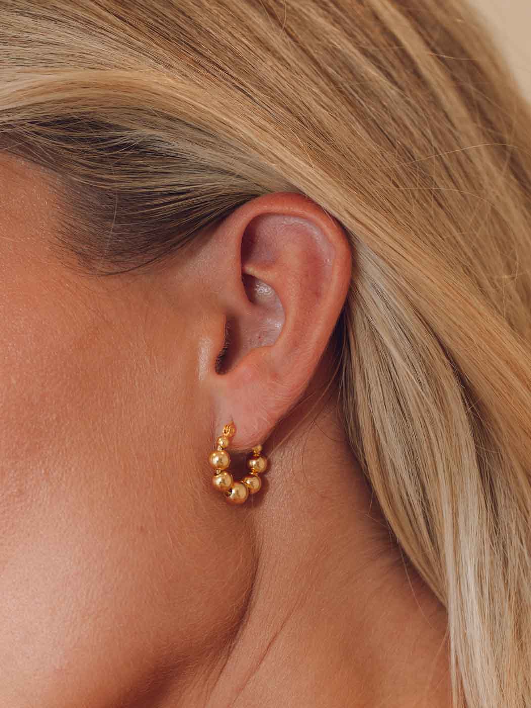A close-up of a model's ear wearing a small gold hoop earring adorned with clustered round gold beads.