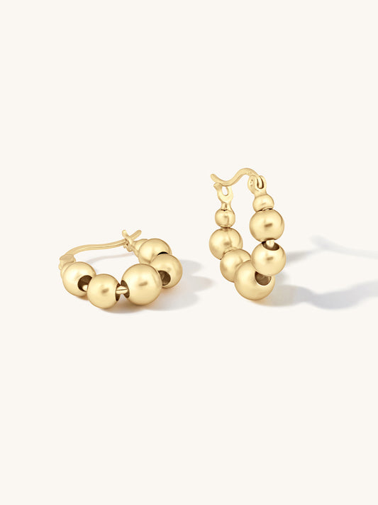Gold hoop earrings with a beaded design, featuring a small cluster of polished gold beads on each hoop, shown on a clean off-white background.