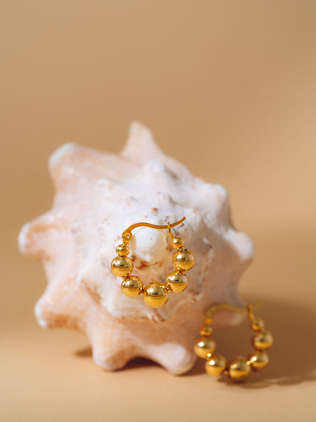 Gold beaded hoop earrings displayed on a white seashell against a warm beige background.