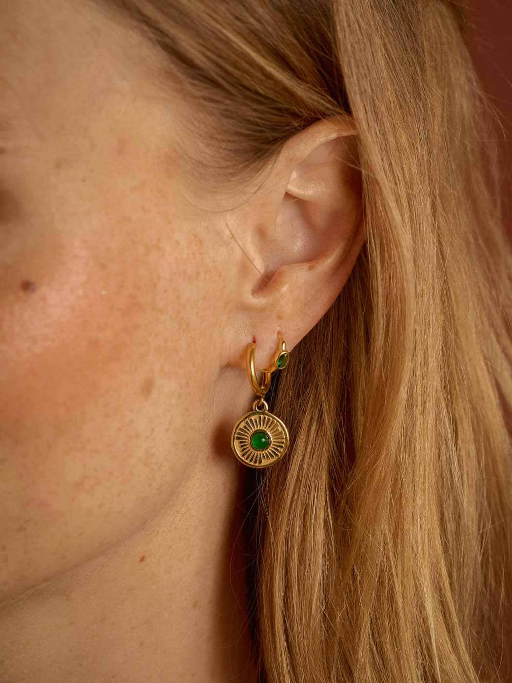 A close-up of a woman's ear wearing a gold huggie hoop earring with a circular sunburst pendant featuring a green center stone.