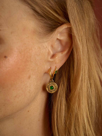 A close-up of a woman's ear wearing a gold huggie hoop earring with a circular sunburst pendant featuring a green center stone.