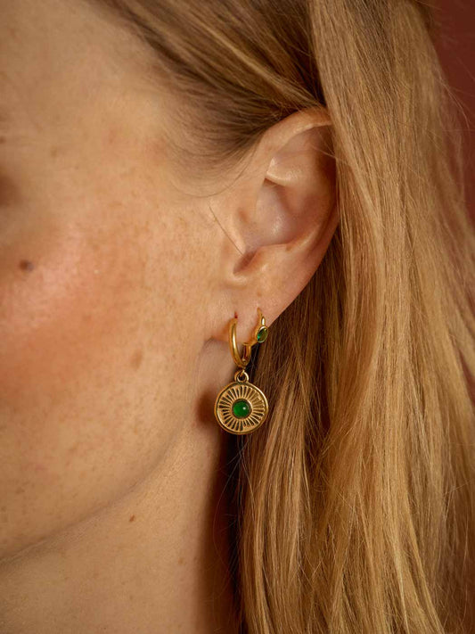 A close-up of a woman's ear wearing a gold huggie hoop earring with a circular sunburst pendant featuring a green center stone.