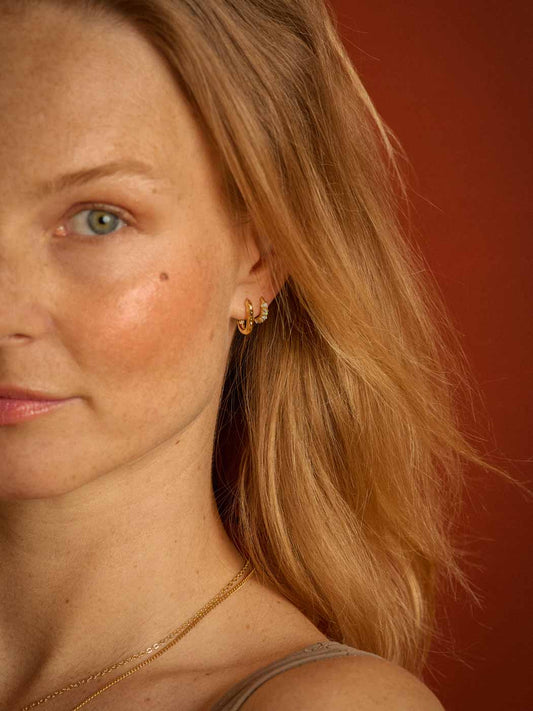 Close-up of a woman with blonde hair wearing a textured oval gold hoop earring and a delicate gold chain necklace against a warm reddish-brown background.