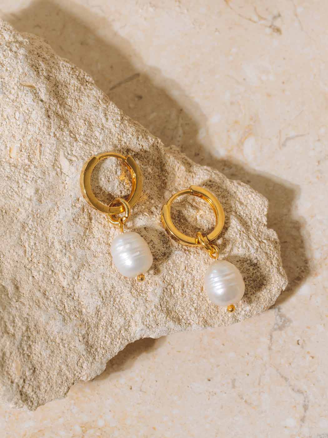 A pair of gold hoop earrings with a dangling white baroque pearl, displayed on a textured beige stone against a matching stone background.