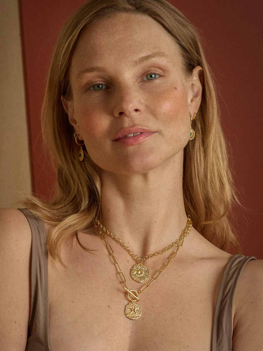 A close-up portrait of a woman wearing layered gold necklaces, including a coin pendant necklace and a chain necklace with a circular medallion, along with matching gold hoop earrings, set against a warm reddish-brown background.