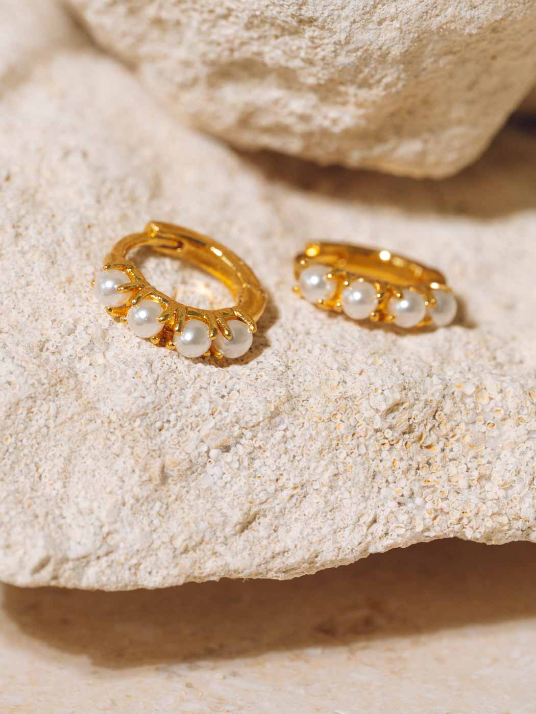 A pair of gold huggie hoop earrings adorned with small white pearls, displayed on a textured beige stone surface.
