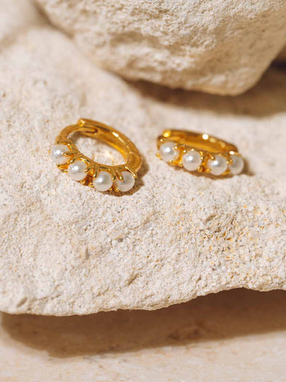A pair of gold huggie hoop earrings adorned with small white pearls, displayed on a textured beige stone surface.