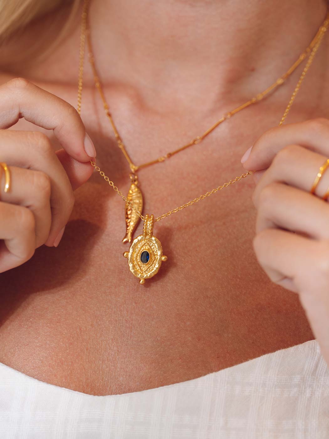 A close-up lifestyle shot of a model wearing a gold layered necklace with a fish and a textured sapphire setting pendant, adjusting the necklace with her hands.