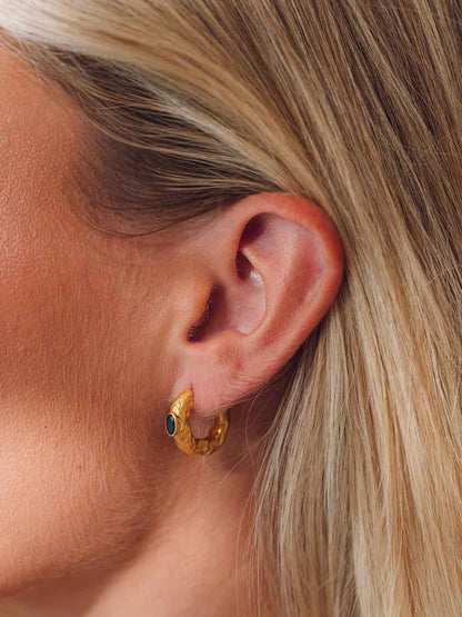 A close-up of a model's ear wearing a textured gold crescent-shaped hoop earring with a small dark green stone accent.