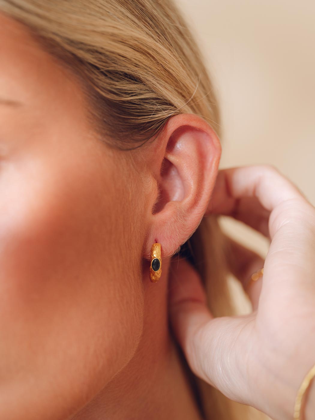A close-up lifestyle shot of a woman wearing a small gold huggie hoop earring with a dark green oval stone, with her hand adjusting the earring on her ear.