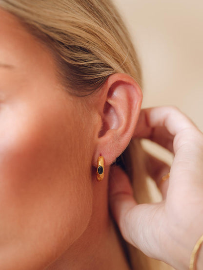 A close-up lifestyle shot of a woman wearing a small gold huggie hoop earring with a dark green oval stone, with her hand adjusting the earring on her ear.