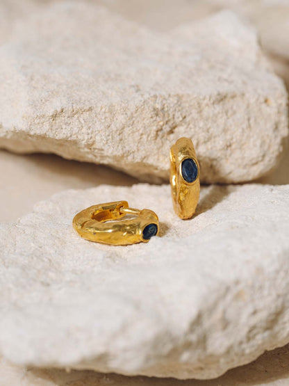 Two gold huggie hoop earrings with dark blue oval gemstones, displayed on textured off-white stones.