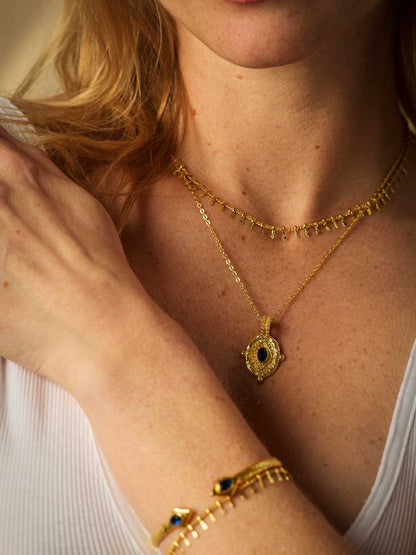 A close-up lifestyle shot of a woman wearing layered gold jewelry, including a gold pendant necklace with a central sapphire stone, a delicate chain necklace, and a gold bracelet with blue and black stones.