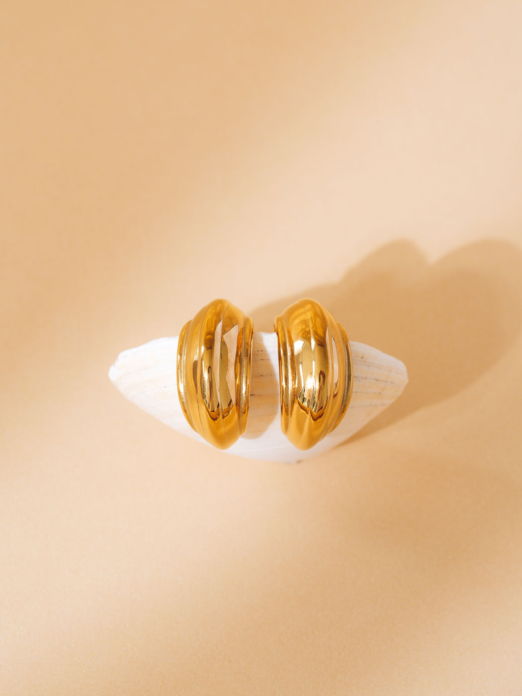 A pair of chunky, polished gold hoop earrings displayed on a small white shell stand against a warm beige background.