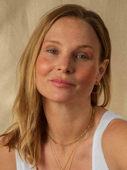 A close-up portrait of a woman with blonde hair wearing layered gold necklaces and gold hoop earrings, against a neutral beige background.