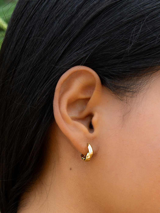 Close-up of an ear wearing a gold twist huggie earring with dark hair and skin in the background