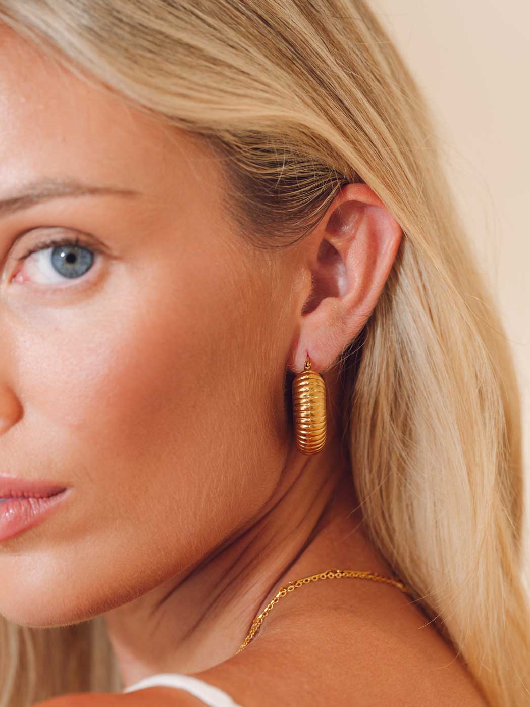 A close-up of a blonde woman wearing a textured gold hoop earring and a delicate gold chain necklace against a soft beige background.
