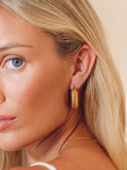 A close-up of a blonde woman wearing a textured gold hoop earring and a delicate gold chain necklace against a soft beige background.