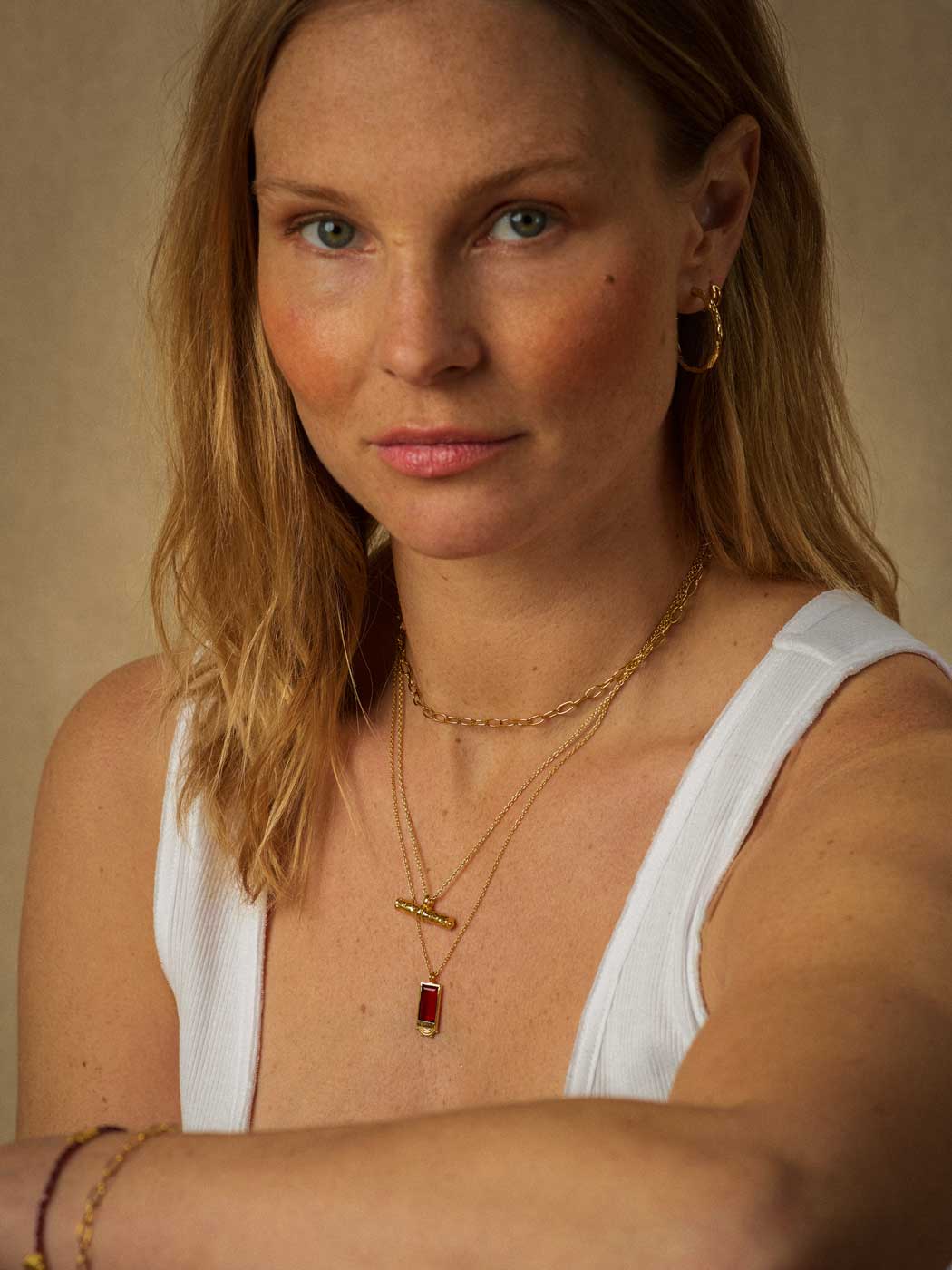 A close-up portrait of a woman wearing layered gold necklaces, including a small rectangular red pendant, a gold hoop earring, and bracelets, paired with a white ribbed tank top against a warm beige background.