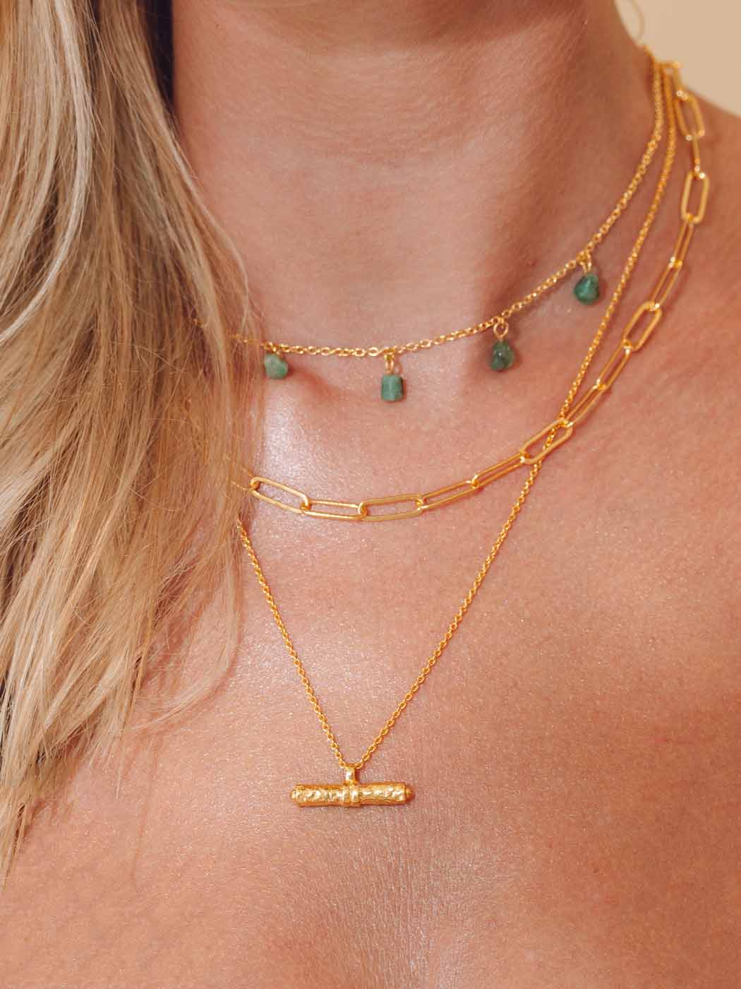 A close-up of a model's neck and chest wearing a layered gold necklace set. The set includes a delicate chain necklace with small green bead pendants, a thicker paperclip chain, and a fine chain with a textured bar pendant.