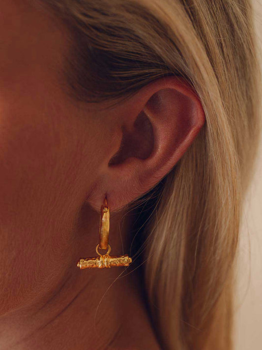 A close-up of a model's ear wearing a textured gold huggie hoop earring with a matching textured gold toggle clasp.