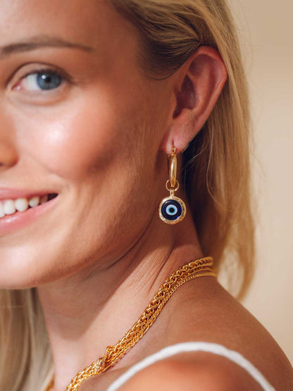 A close-up lifestyle shot of a woman wearing a gold huggie hoop earring with a circular evil eye charm, paired with a gold chain necklace.