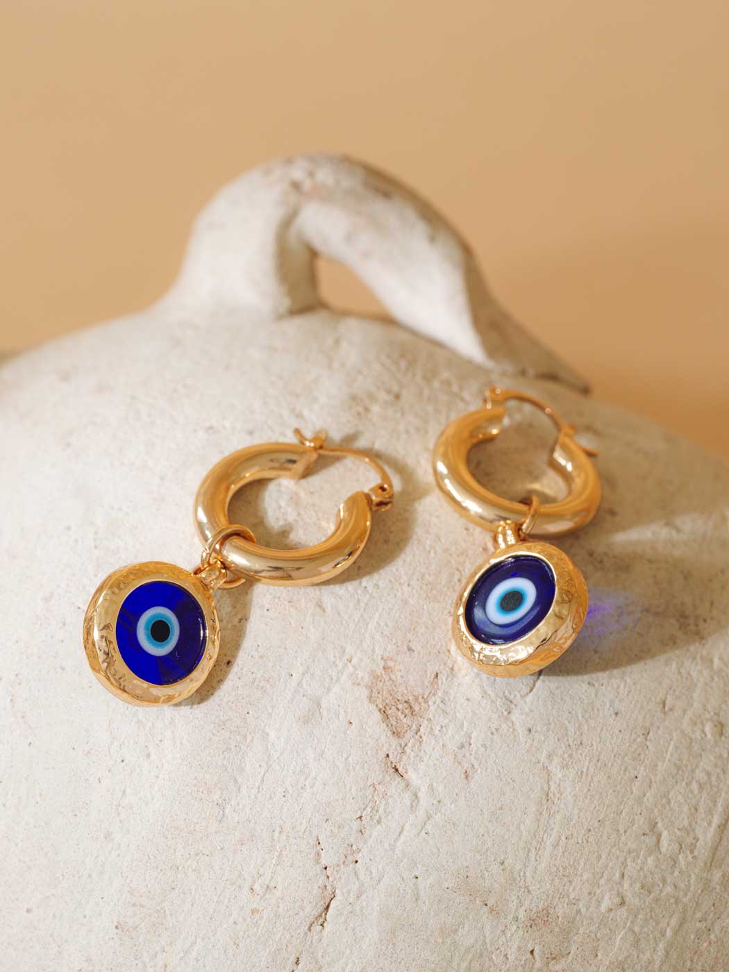 A pair of gold hoop earrings with circular evil eye charms, displayed on a textured beige stone surface against a warm beige background.