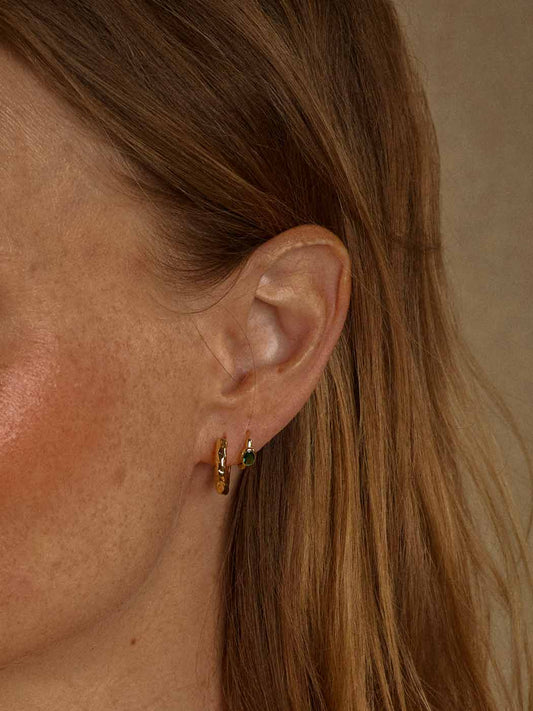 Close-up of a woman's ear wearing a pair of small gold hoop earrings with tiny green and white gemstone accents, against a neutral beige background.