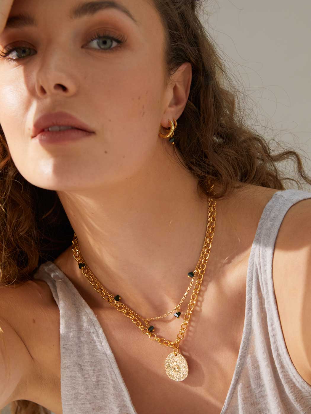A close-up lifestyle shot of a woman wearing a layered gold necklace with a textured oval pendant and small black bead accents, along with a gold hoop earring.
