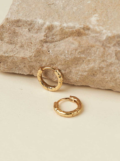 Two small textured gold huggie hoop earrings displayed on a light cream surface with a large beige stone prop in the background.