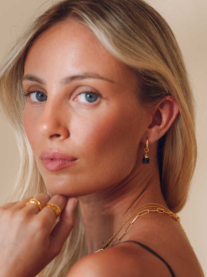 A close-up portrait of a blonde woman wearing a small gold huggie hoop earring with a dark rectangular stone, along with gold rings and layered gold necklaces, against a warm beige background.