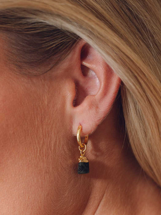 A close-up of a model's ear wearing a small gold huggie hoop earring with a dangling dark green faceted gemstone.