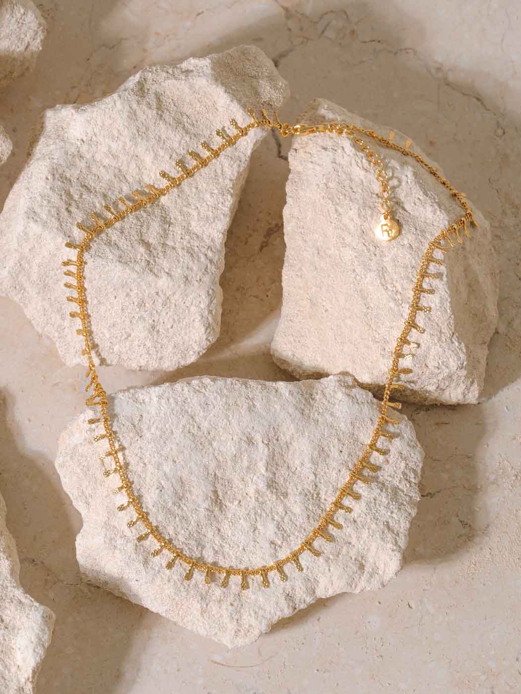 A delicate gold chain necklace with small dangling charms, draped over rough, light beige stones against a matching stone background.