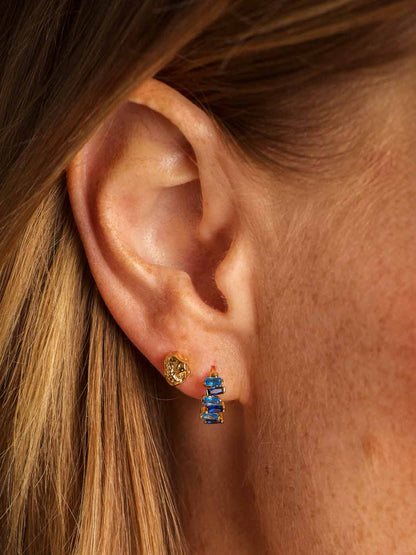 Close-up of a person's ear wearing a small gold stud earring with a stacked blue crystal cluster huggie