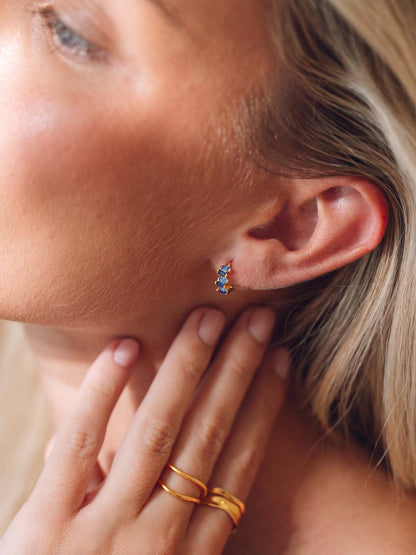 A close-up lifestyle shot of a woman wearing a small blue gemstones huggie, with her hand resting on her neck.