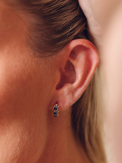 A close-up of a woman's ear wearing a small gold stud earring with three stacked rectangular blue gemstones.