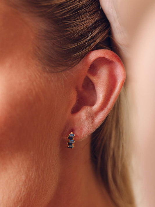 A close-up of a woman's ear wearing a small gold stud earring with three stacked rectangular blue gemstones.