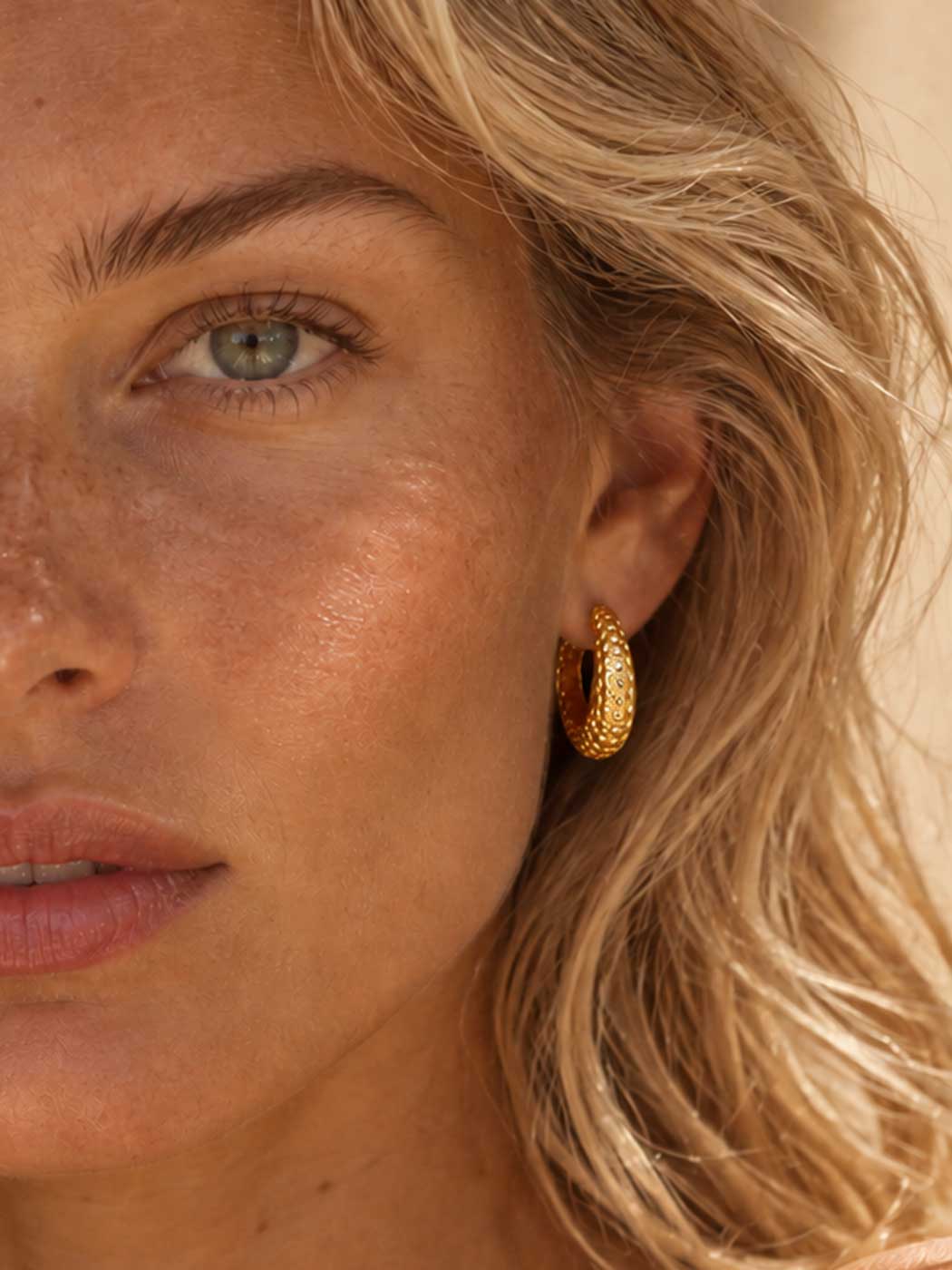 Close-up of a woman wearing a textured gold hoop earring, showing the earring on her ear against her blonde hair and warm skin tones.