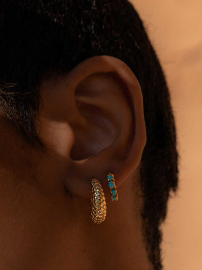 A close-up of a person's ear wearing two gold hoop earrings, one larger textured gold hoop and one smaller gold hoop with small turquoise blue stones, against a warm beige background.