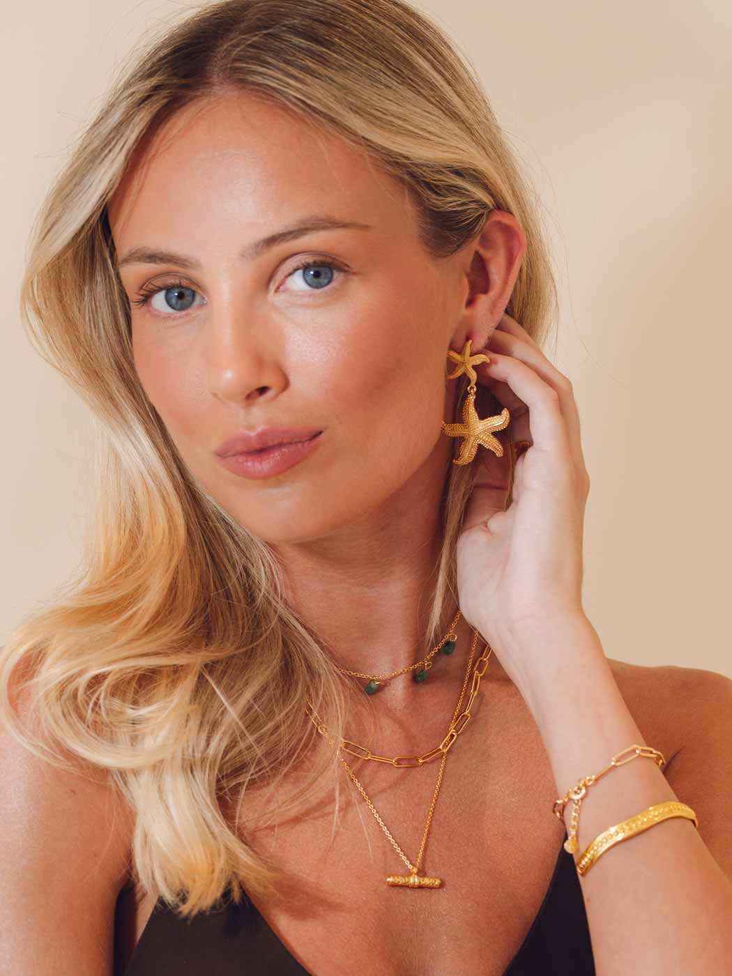 Woman wearing gold jewelry including earrings, necklace, and bracelet against a beige background