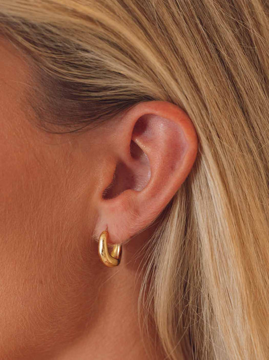 A close-up of a model's ear wearing a small, polished gold hoop earring.