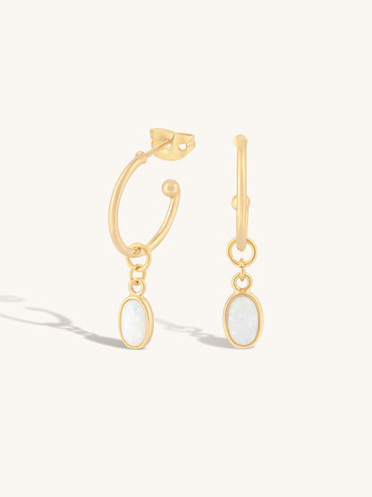 A pair of gold hoop earrings with oval white opal-like dangles, shown on a clean off-white background with soft shadows.
